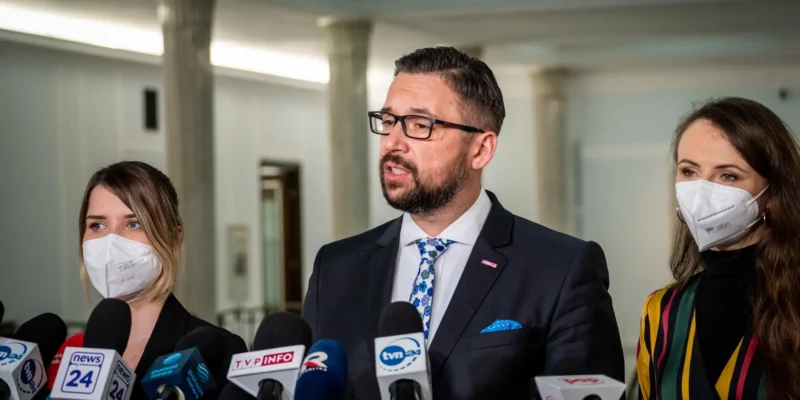 Mężczyzna w garniturze i okularach przemawia do mediów podczas konferencji prasowej, otoczony przez dwie kobiety w maseczkach.