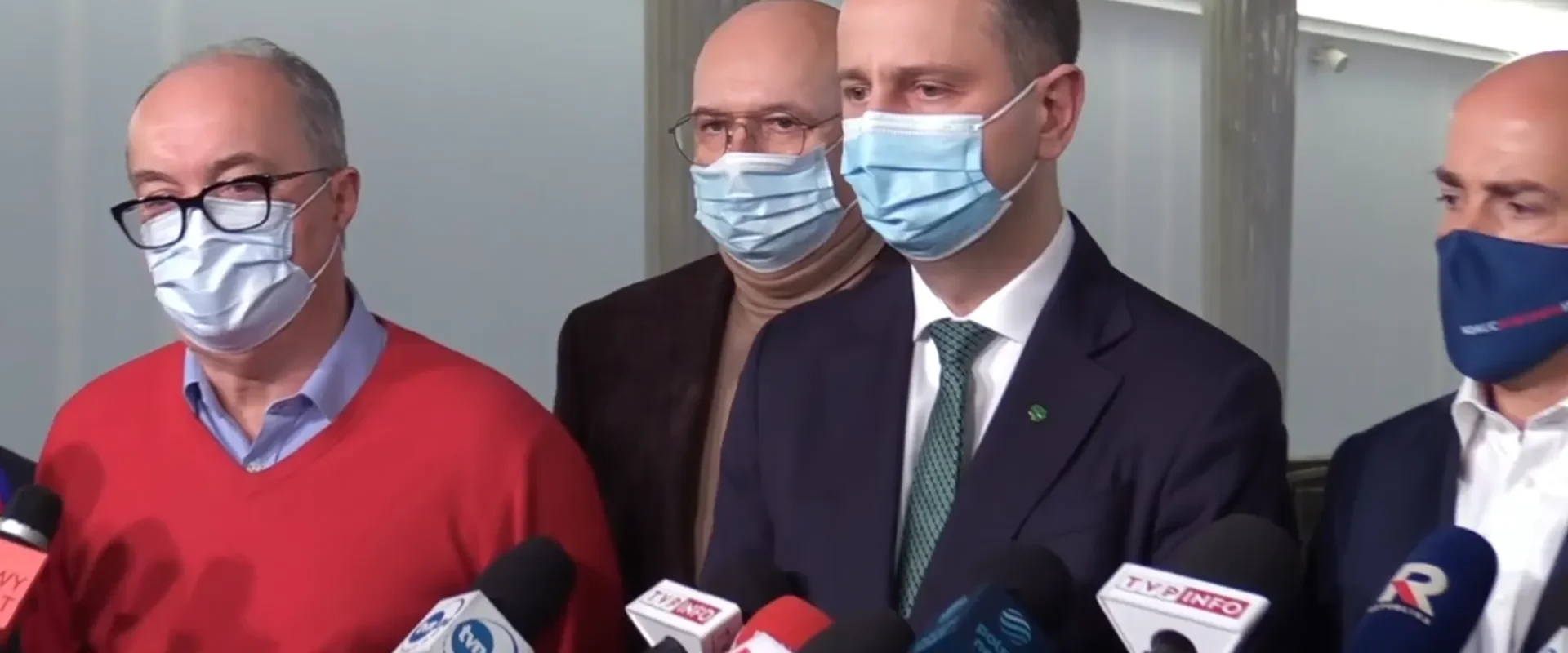 Grupa mężczyzn w maseczkach stoi na konferencji prasowej, udzielając wypowiedzi mediom przed licznymi mikrofonami.