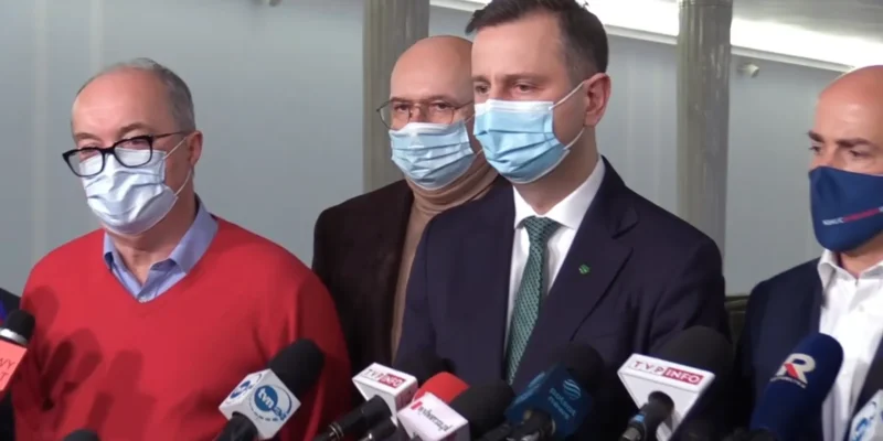 Grupa mężczyzn w maseczkach stoi na konferencji prasowej, udzielając wypowiedzi mediom przed licznymi mikrofonami.