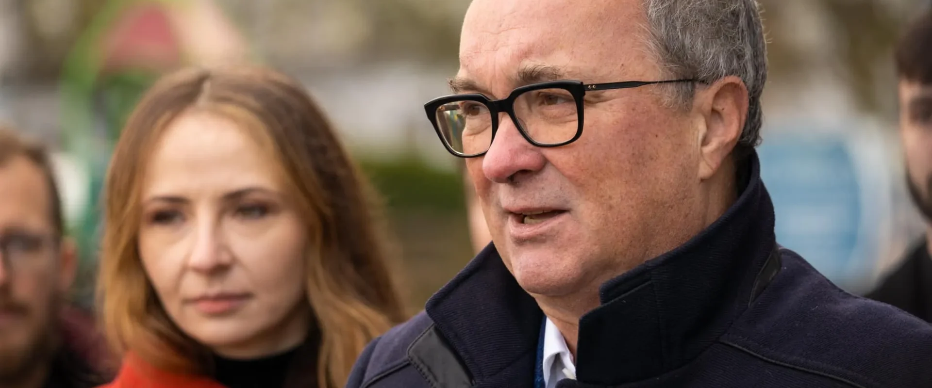 Mężczyzna w czarnych okularach i ciemnym płaszczu wypowiada się do mikrofonu podczas konferencji, w tle stoi kobieta z długimi jasnymi włosami.