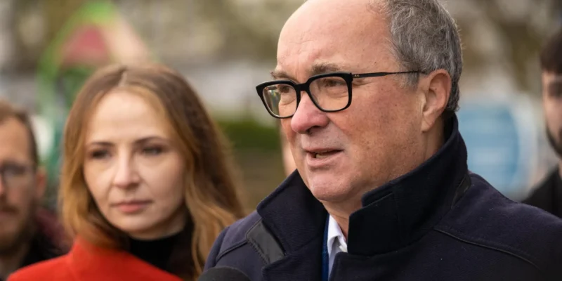 Mężczyzna w czarnych okularach i ciemnym płaszczu wypowiada się do mikrofonu podczas konferencji, w tle stoi kobieta z długimi jasnymi włosami.