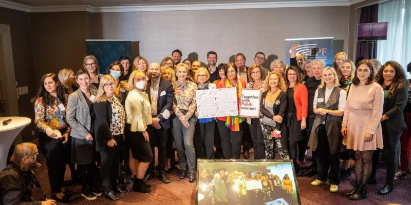 Duża grupa osób stojących razem w sali konferencyjnej podczas wydarzenia manifestu Warszawskiego, na środku trzyma podpisany dokument i plakat „Together with Polish women”, w tle widoczne banery organizacyjne.