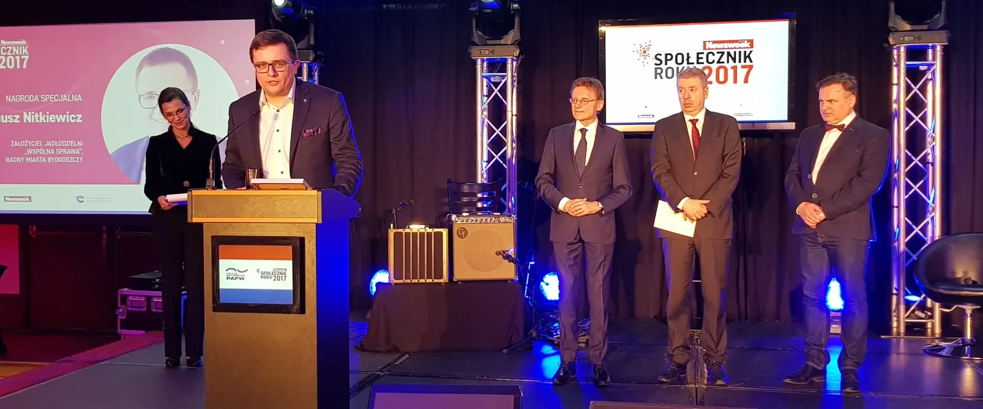 Mężczyzna przy mównicy odbiera nagrodę specjalną podczas gali "Społecznik Roku 2017", obok stoją trzy osoby i widoczne są ekrany z nazwą wydarzenia.