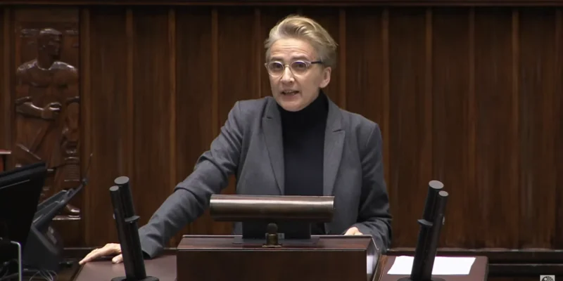 Na zdjęciu przedstawiona jest kobieta w szarej marynarce, mówiąca z mównicy w sali obrad, z drewnianym tłem i mikrofonami w zasięgu ręki.