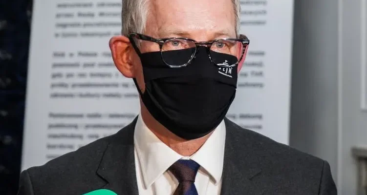 Mężczyzna w garniturze, noszący czarną maseczkę i okulary, stoi przy mikrofonach podczas konferencji prasowej.