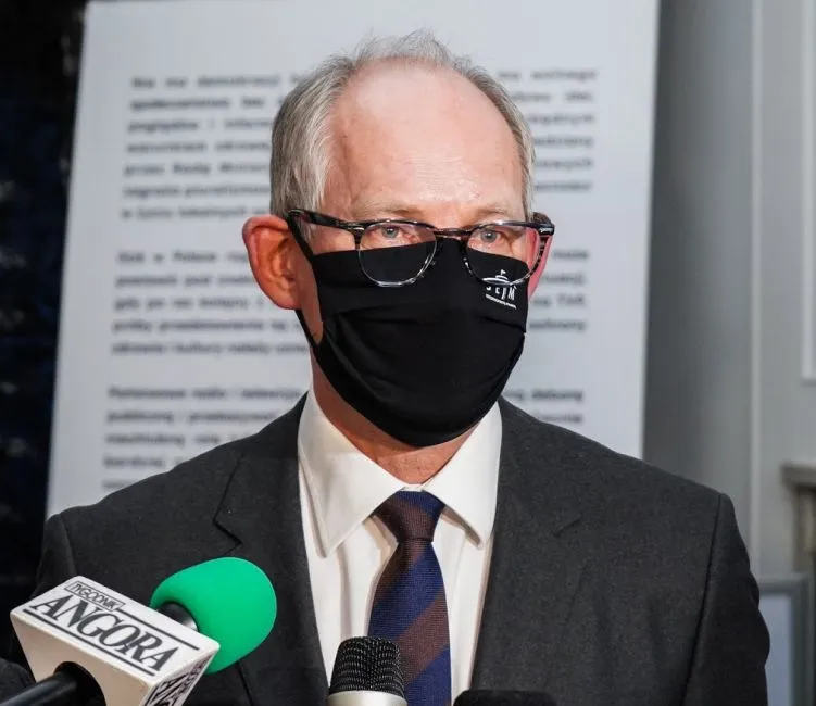 Mężczyzna w garniturze, noszący czarną maseczkę i okulary, stoi przy mikrofonach podczas konferencji prasowej.