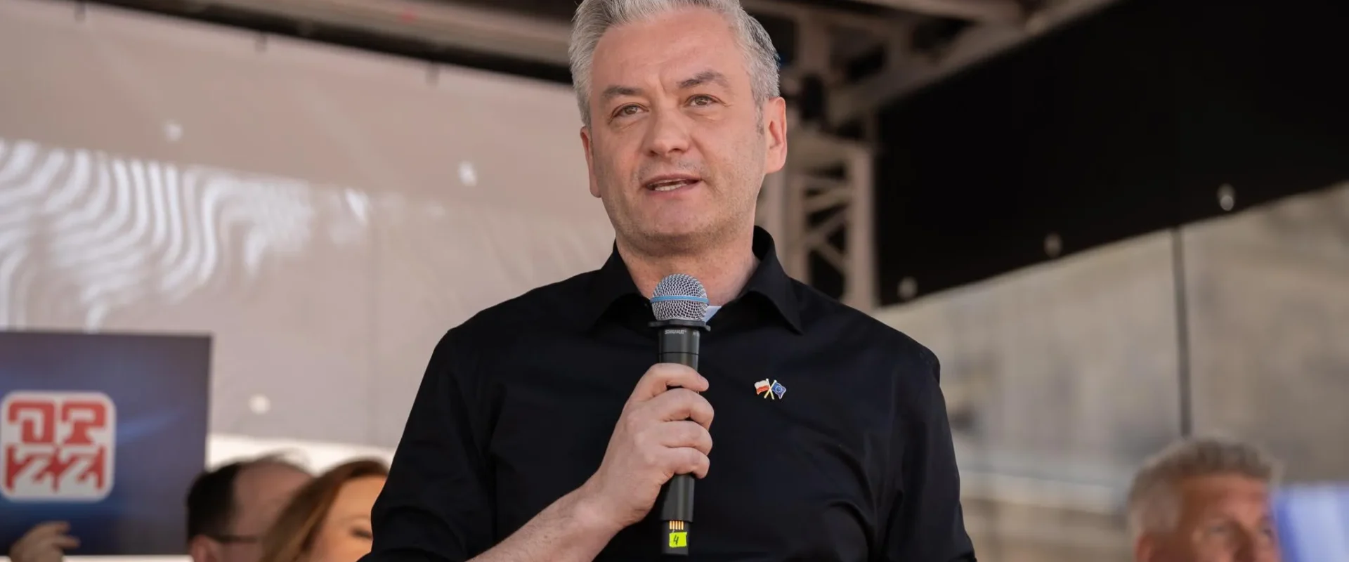 Mężczyzna w czarnej koszuli trzyma mikrofon i przemawia na scenie podczas wydarzenia.