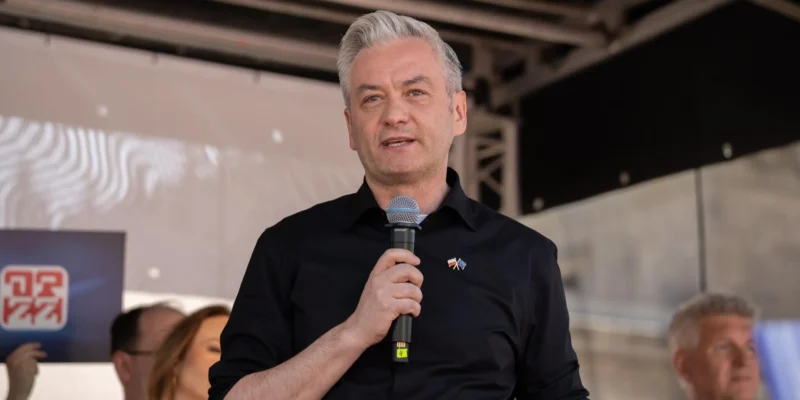 Mężczyzna w czarnej koszuli trzyma mikrofon i przemawia na scenie podczas wydarzenia.