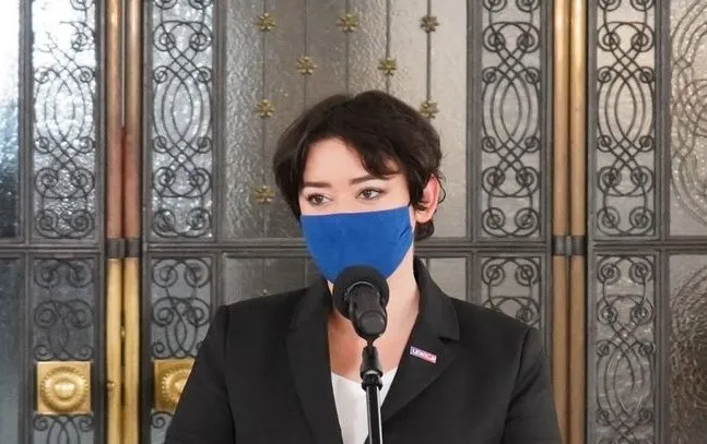 Osoba ubrana w czarny żakiet stoi przy mikrofonie, nosi niebieską maseczkę, a w tle widać ozdobne metalowe drzwi.