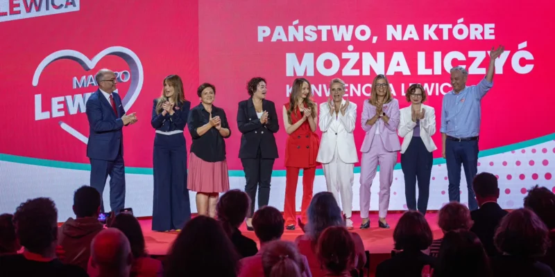 Grupa polityków stoi na scenie podczas wydarzenia, klaszcze i uśmiecha się, na tle dużego różowo-czerwonego baneru z napisem „PAŃSTWO, NA KTÓRE MOŻNA LICZYĆ” i logiem Lewicy.