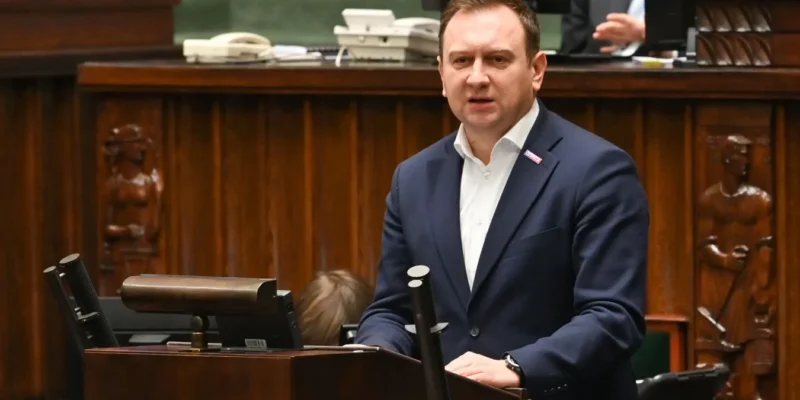 Mężczyzna w granatowym garniturze przemawia z mównicy podczas debaty budżetowej w sali obrad, na tle drewnianych paneli.