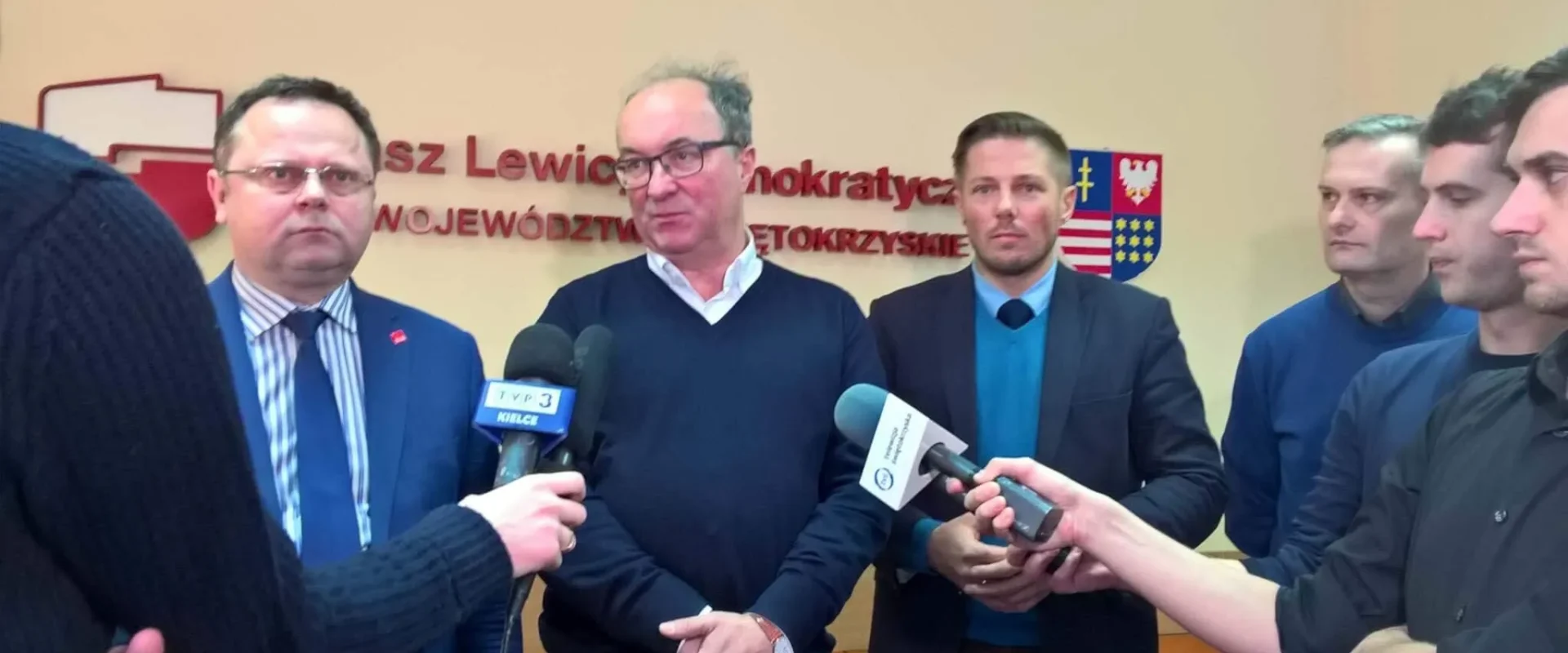 Kilku mężczyzn stoi na konferencji prasowej przed mikrofonami, z napisem i herbem na ścianie za nimi.