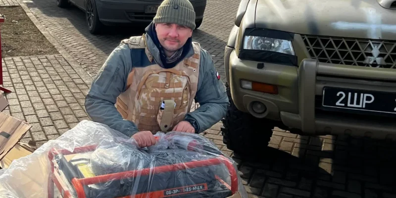 Mężczyzna w kamizelce ochronnej siedzi na opakowaniach, trzymając generator w foliowym pakowaniu, obok pojazdów zaparkowanych na utwardzonej drodze.