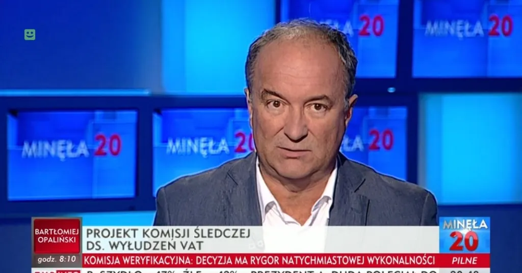 Mężczyzna w marynarce przemawia w studiu telewizyjnym z wieloma ekranami „Minęła 20” w tle i paskiem informacyjnym o komisji śledczej ds. wyłudzeń VAT.