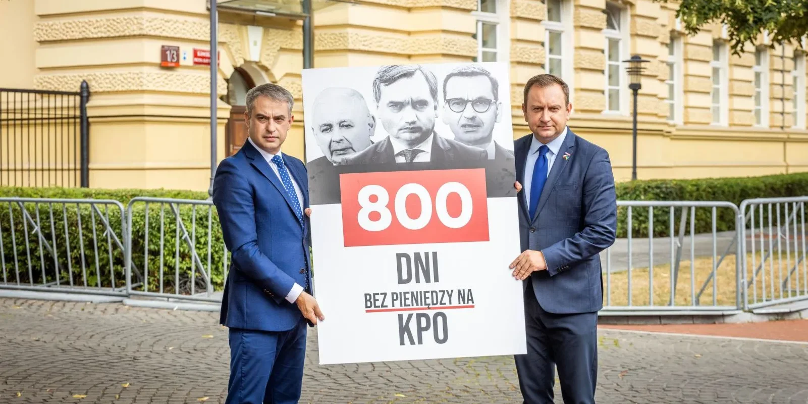 Dwóch mężczyzn w garniturach stoi na chodniku przed żółtym budynkiem, trzymając duży plakat z napisem „800 dni bez pieniędzy na KPO” i czarno-białymi wizerunkami polityków na górze.