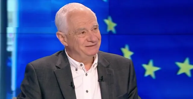 Starszy mężczyzna w ciemnym garniturze i białej koszuli uśmiecha się podczas wywiadu telewizyjnego, na niebieskim tle widoczne żółte gwiazdy symbolizujące Unię Europejską.