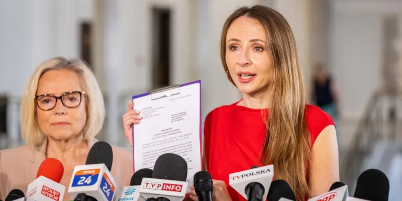 Dwie kobiety podczas konferencji prasowej, z których jedna trzyma dokument na kartce, otoczone licznymi mikrofonami mediów.