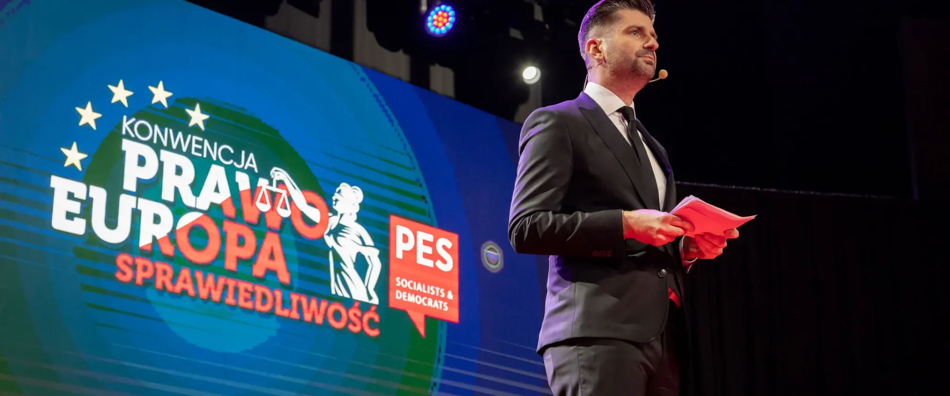 Mężczyzna w garniturze przemawia na tle kolorowego ekranu z napisem "Konwencja Prawo Europa Sprawiedliwość".