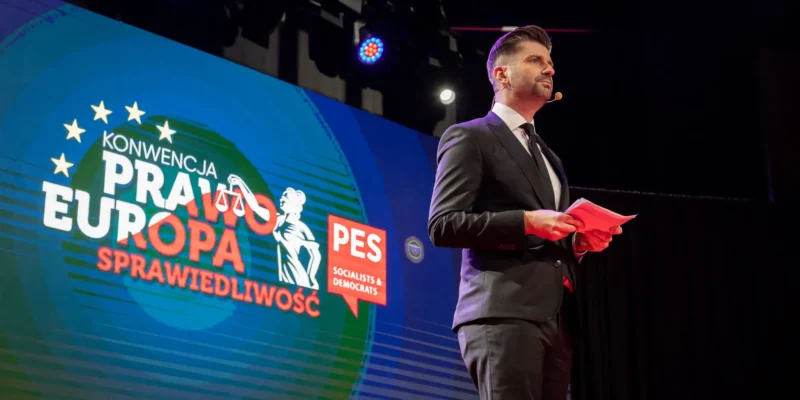 Mężczyzna w garniturze przemawia na tle kolorowego ekranu z napisem "Konwencja Prawo Europa Sprawiedliwość".