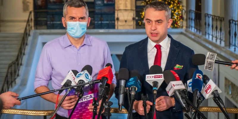 Dwóch mężczyzn w garniturach stoi za szeregiem mikrofonów podczas konferencji prasowej w eleganckim holu z choinką w tle; z lewej – w fioletowej koszuli i maseczce, z prawej – w granatowym garniturze z czerwonym krawatem.