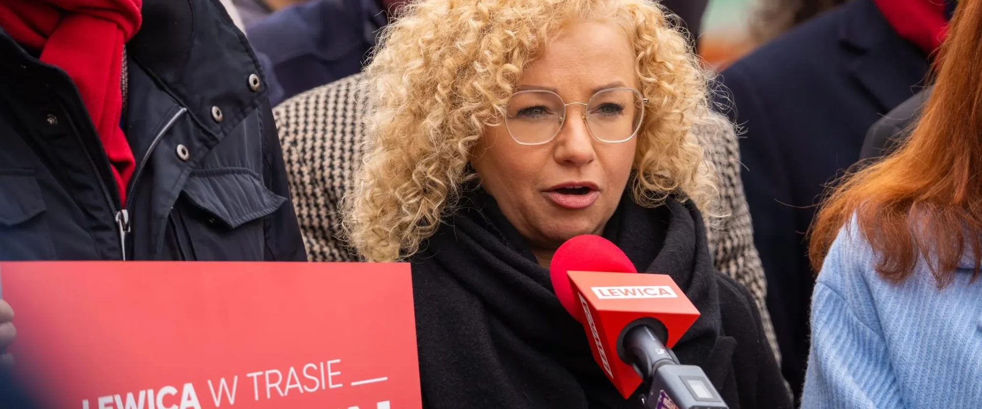 Na zdjęciu przedstawiona jest kobieta z kręconymi włosami, mówiąca do mikrofonu podczas wydarzenia związanego z Lewicą w Szczecinie, trzymająca czerwoną planszę z napisem "LEWICA W TRASIE".