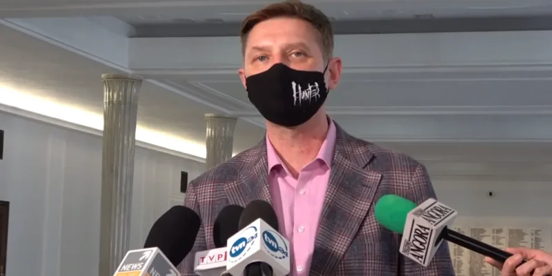 Mężczyzna w różowej koszuli i kraciastej marynarce, noszący czarną maseczkę z napisem Hunter, udziela konferencji prasowej przed zestawem mikrofonów.