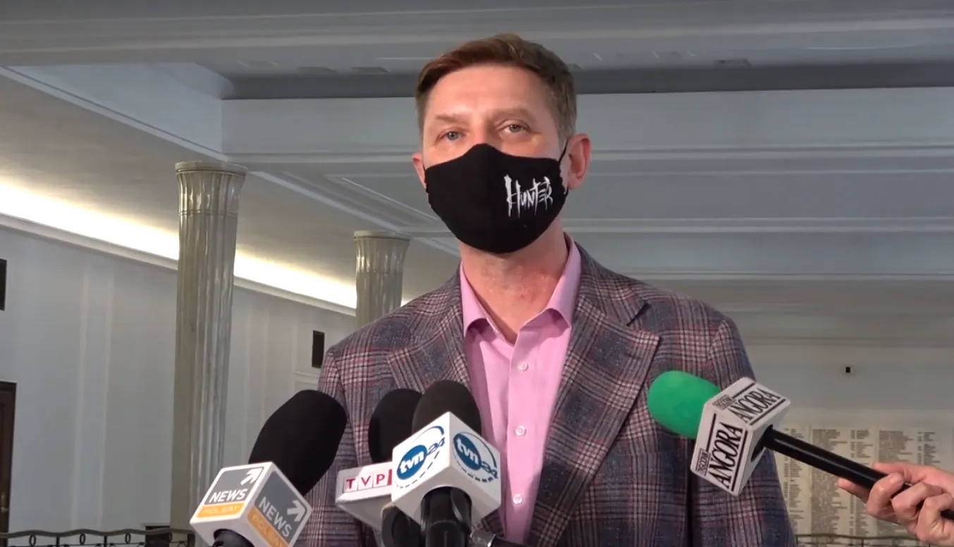 Mężczyzna w różowej koszuli i kraciastej marynarce, noszący czarną maseczkę z napisem Hunter, udziela konferencji prasowej przed zestawem mikrofonów.