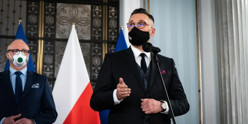 Mężczyzna w czarnym garniturze i maseczce przemawia do mikrofonu, stojąc obok drugiego mężczyzny w garniturze; w tle widoczne flagi Polski i Unii podczas wydarzenia „Wspólnie dla Polski”.