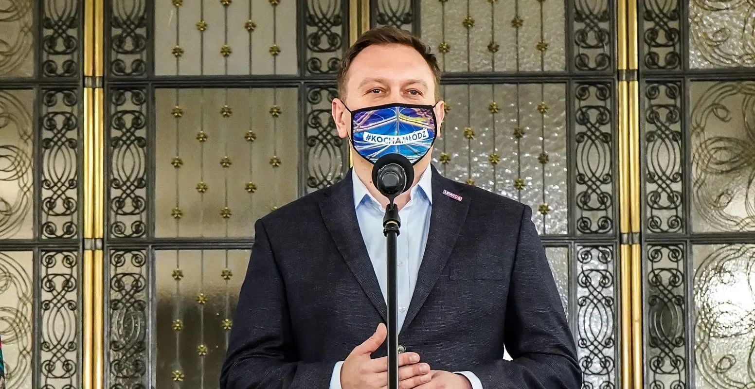 Mężczyzna w garniturze stoi przy mikrofonie podczas konferencji prasowej, nosi maseczkę z napisem #KOCHAMŁÓDŹ, a w tle widoczne są ozdobne metalowe drzwi.