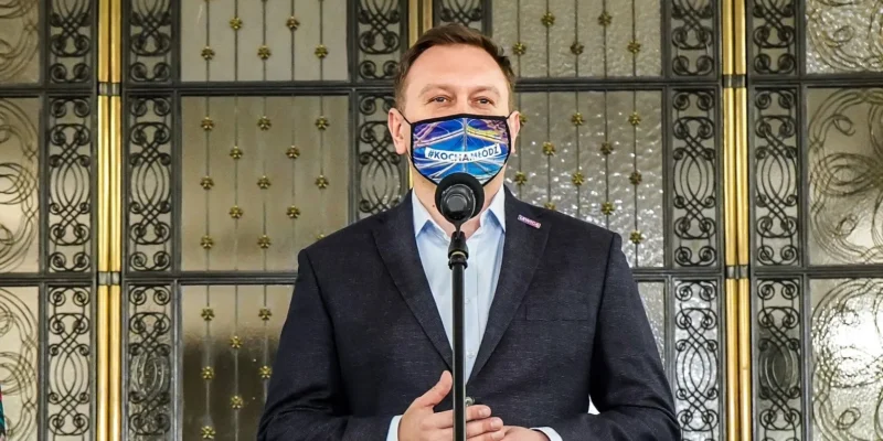 Mężczyzna w garniturze stoi przy mikrofonie podczas konferencji prasowej, nosi maseczkę z napisem #KOCHAMŁÓDŹ, a w tle widoczne są ozdobne metalowe drzwi.