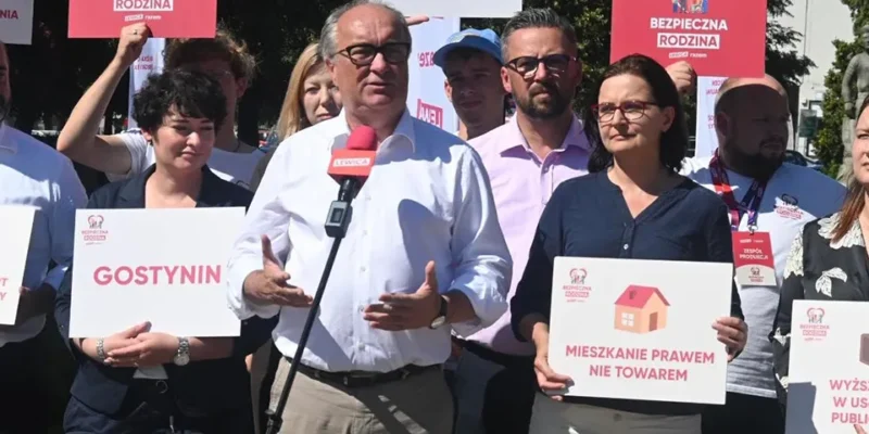 Na zdjęciu przedstawiono grupę osób uczestniczących w demonstracji, trzymających transparenty z hasłami społecznymi, a w centrum występuje mężczyzna mówiący do mikrofonu.