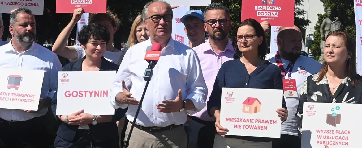 Na zdjęciu przedstawiono grupę osób uczestniczących w demonstracji, trzymających transparenty z hasłami społecznymi, a w centrum występuje mężczyzna mówiący do mikrofonu.