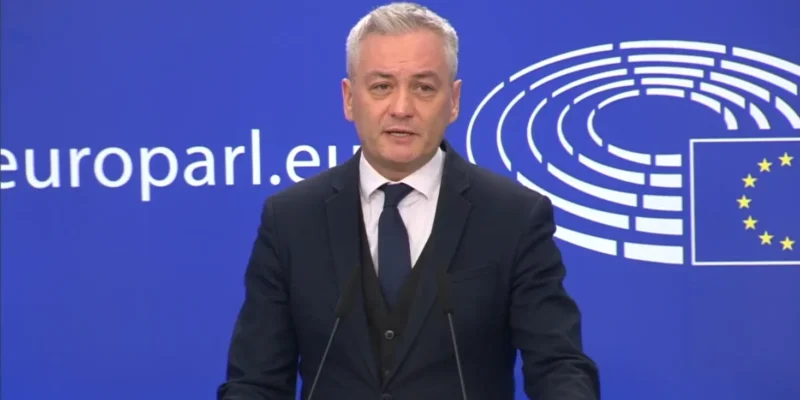 Mężczyzna w eleganckim garniturze przemawia podczas konferencji prasowej przed tłem z logo Parlamentu Europejskiego.