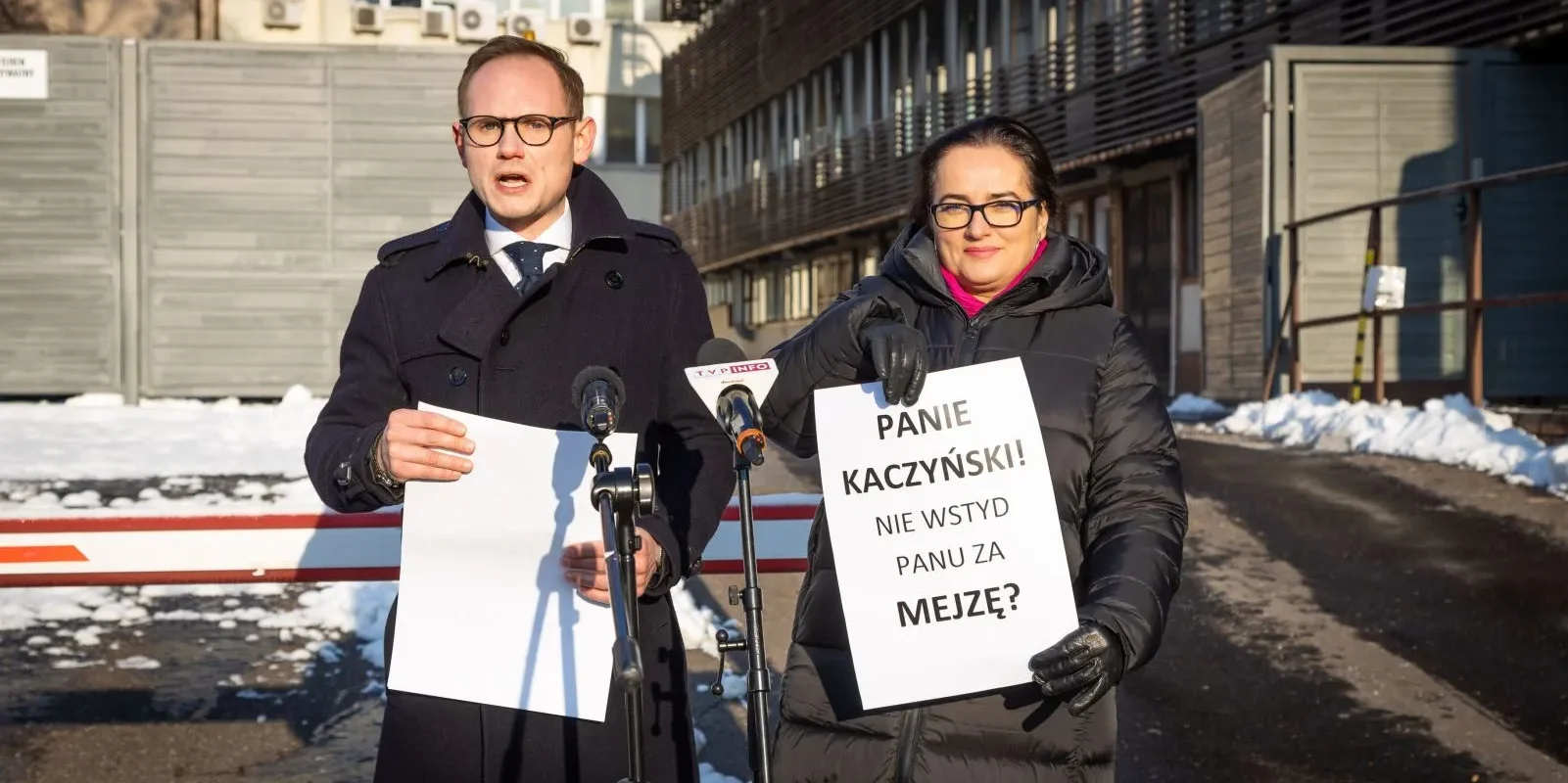 Na zdjęciu widać dwóch mówców podczas konferencji prasowej, trzymających tablice z napisami, stojących przed budynkiem w zimowej scenerii.
