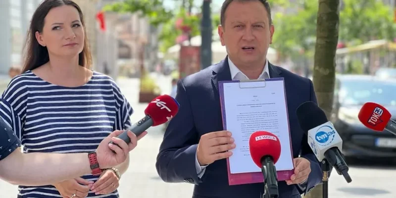 Na ulicy dwie osoby prowadzą konferencję prasową: mężczyzna w garniturze trzyma dokument w fioletowej teczce, a obok niego stoi kobieta; przed nimi ustawione są mikrofony mediów.