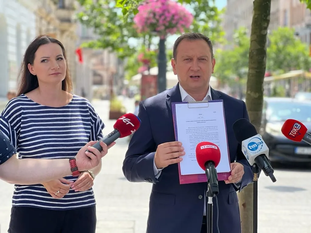 Na ulicy dwie osoby prowadzą konferencję prasową: mężczyzna w garniturze trzyma dokument w fioletowej teczce, a obok niego stoi kobieta; przed nimi ustawione są mikrofony mediów.