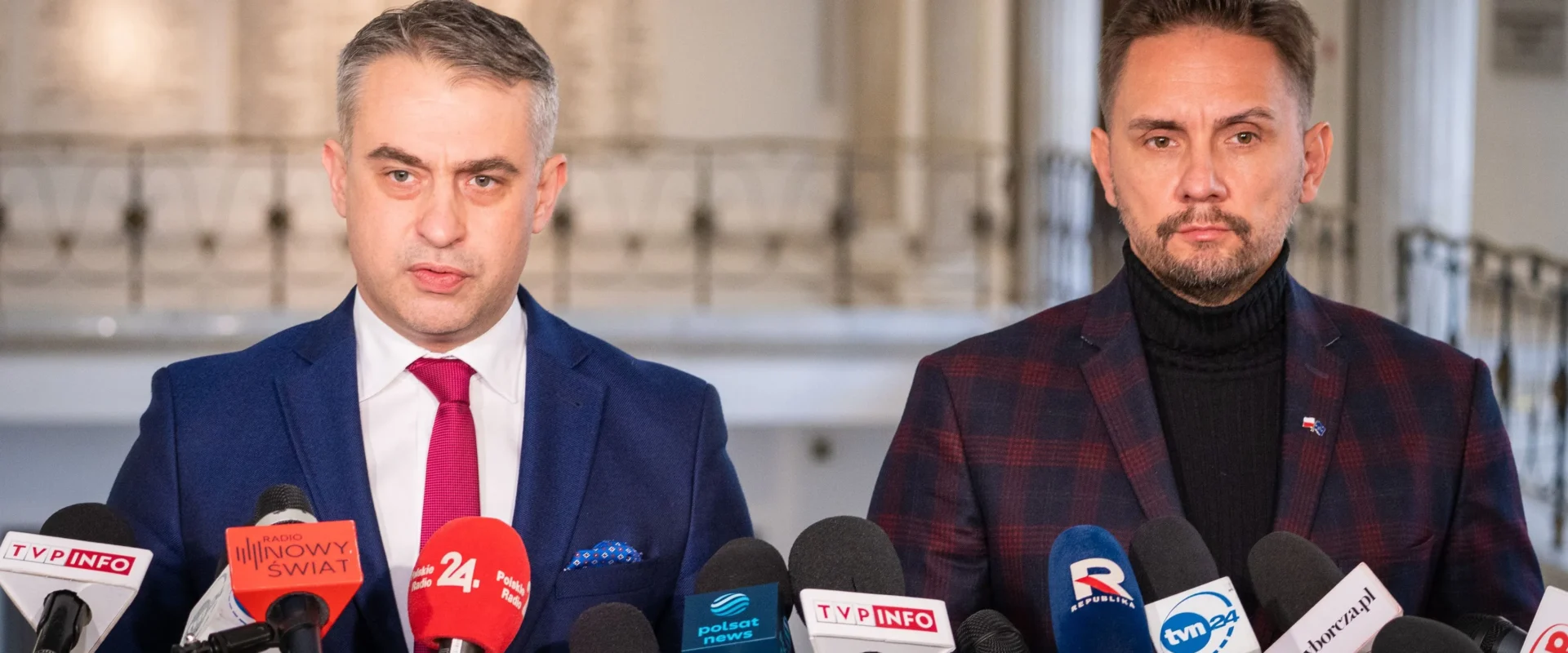 Dwóch mężczyzn w garniturach stoi przed mikrofonami na konferencji prasowej w przestrzeni publicznej.