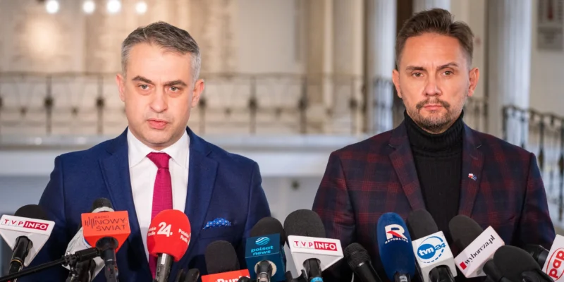 Dwóch mężczyzn w garniturach stoi przed mikrofonami na konferencji prasowej w przestrzeni publicznej.