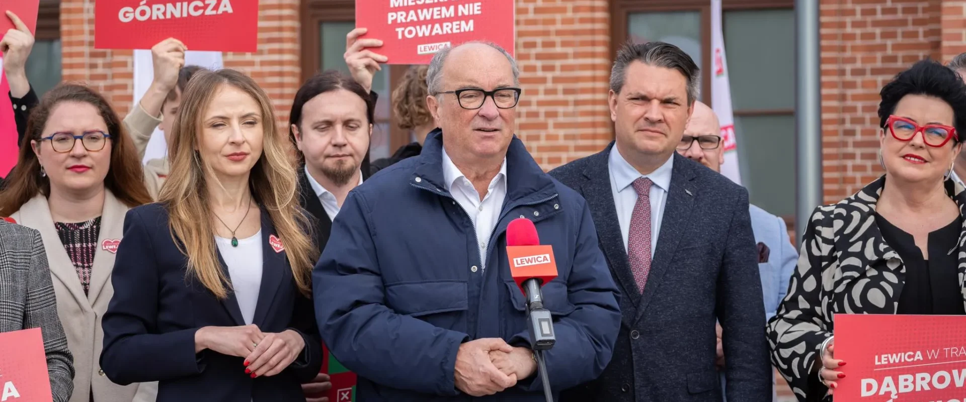 Grupa polityków Lewicy na zewnątrz podczas konferencji prasowej, lider przemawia do mikrofonu z logiem Lewica, wokół niego stoją czerwone plakaty z hasłami wyborczymi, akcentujące postulat 3% PKB na obronność.