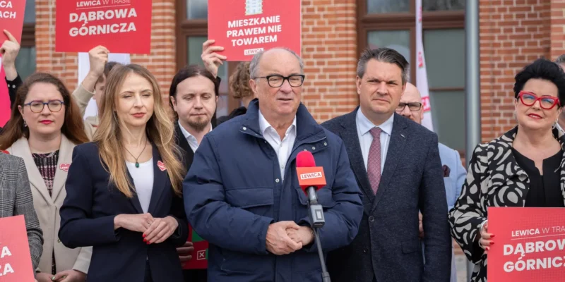 Grupa polityków Lewicy na zewnątrz podczas konferencji prasowej, lider przemawia do mikrofonu z logiem Lewica, wokół niego stoją czerwone plakaty z hasłami wyborczymi, akcentujące postulat 3% PKB na obronność.