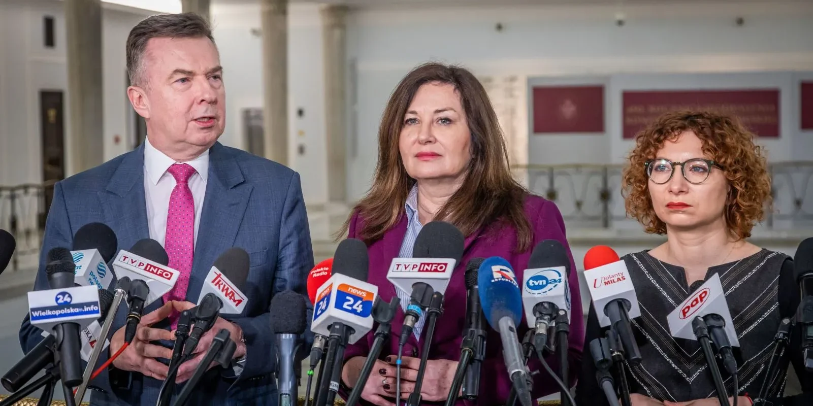 Trzech polityków stoi przy zestawie mikrofonów podczas konferencji prasowej w holu, omawiając nowelizację ustawy wiatrakowej i program Koryto Plus.