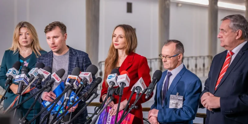 Na zdjęciu widać grupę osób stojących przed mikrofonami podczas konferencji prasowej.