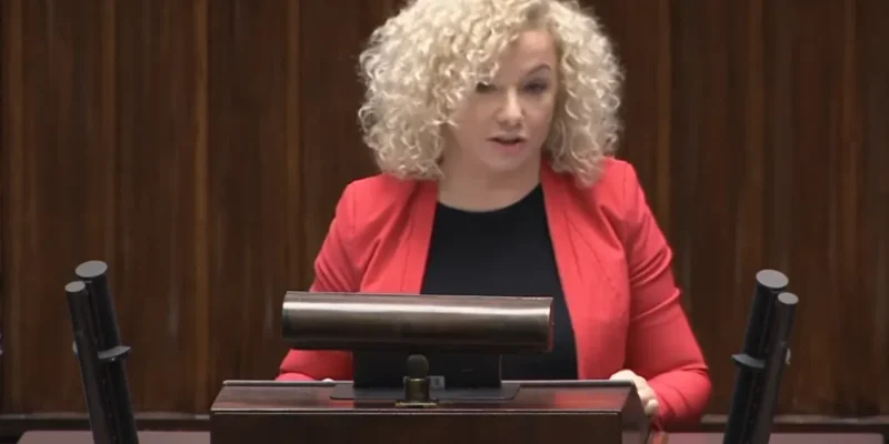 Kobieta o kręconych blond włosach w czerwonej marynarce stoi za podium i przemawia do publiczności.