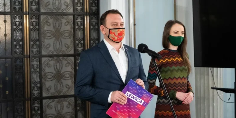 Dwóch ludzi w maseczkach stoi przy mikrofonie podczas konferencji; mężczyzna w granatowym garniturze trzyma różowo-fioletową ulotkę z napisem LEWICA, obok stoi kobieta w kolorowej wzorzystej sukience.