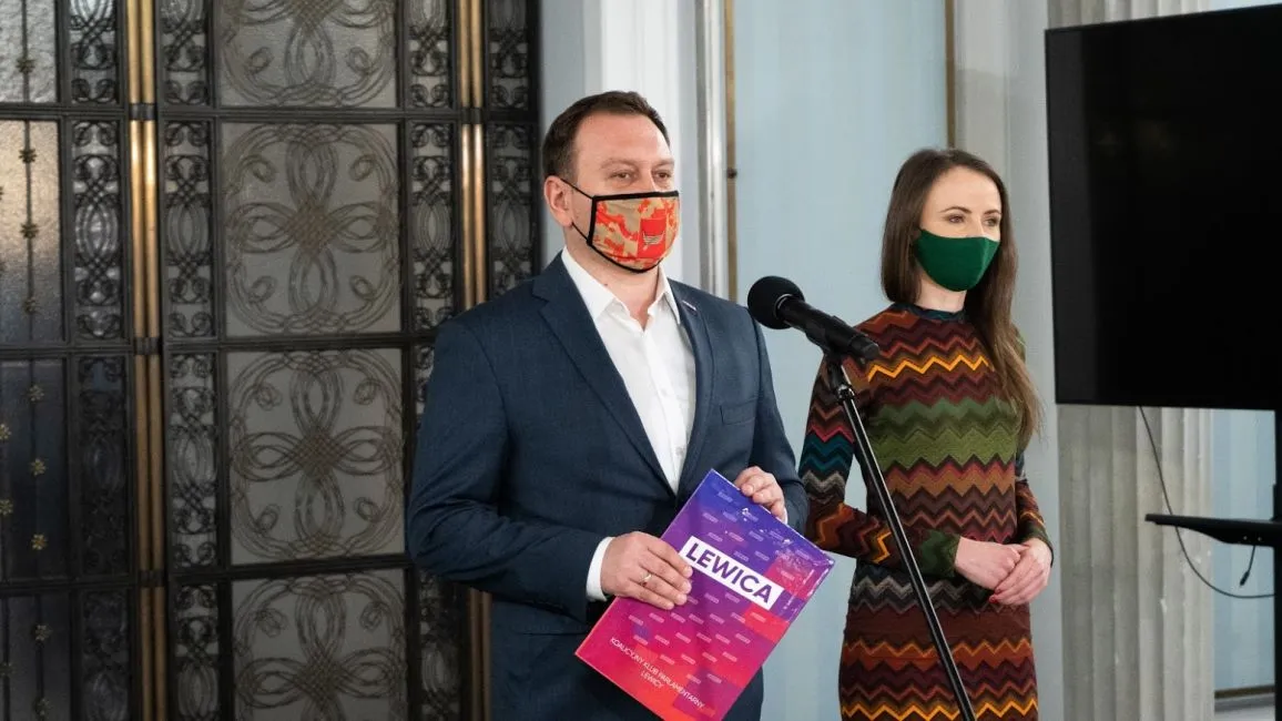 Dwóch ludzi w maseczkach stoi przy mikrofonie podczas konferencji; mężczyzna w granatowym garniturze trzyma różowo-fioletową ulotkę z napisem LEWICA, obok stoi kobieta w kolorowej wzorzystej sukience.