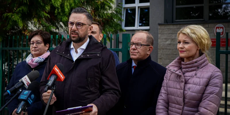 Grupa lokalnych działaczy i polityków wygłasza konferencję prasową przed budynkiem, broniąc społeczności lokalnych i podkreślając hasło „Nie ma demokracji bez samorządności”.