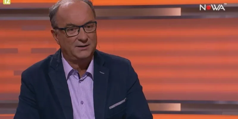 Mężczyzna w marynarce i okularach siedzi w studiu telewizyjnym na pomarańczowym tle podczas wywiadu.