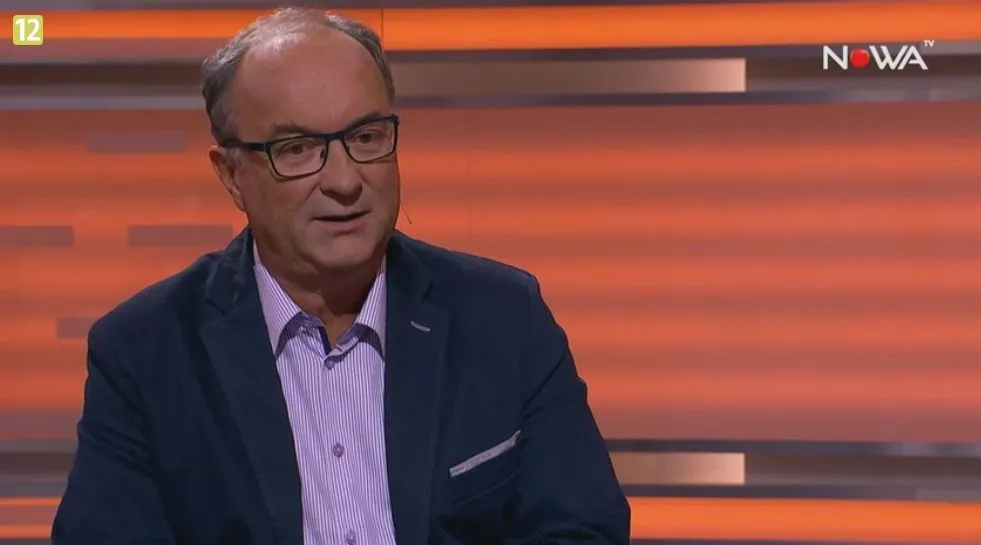 Mężczyzna w marynarce i okularach siedzi w studiu telewizyjnym na pomarańczowym tle podczas wywiadu.