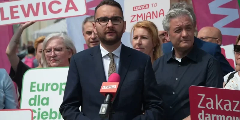 Polityk Kowal w garniturze przemawia na wiecu Lewicy, przedstawiając propozycję powołania Europejskiej Unii Energetycznej; w tle widoczni demonstranci i kolorowe banery.