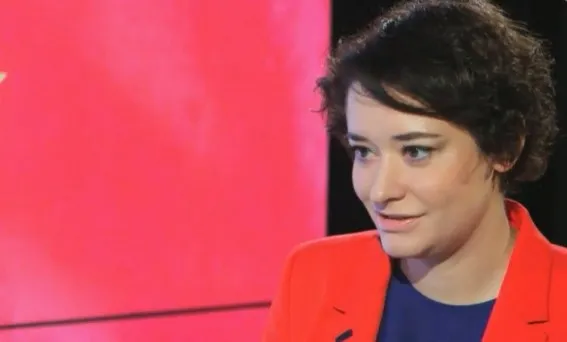 Kobieta w czerwonej marynarce siedzi w studiu telewizyjnym i uśmiecha się, patrząc w bok.